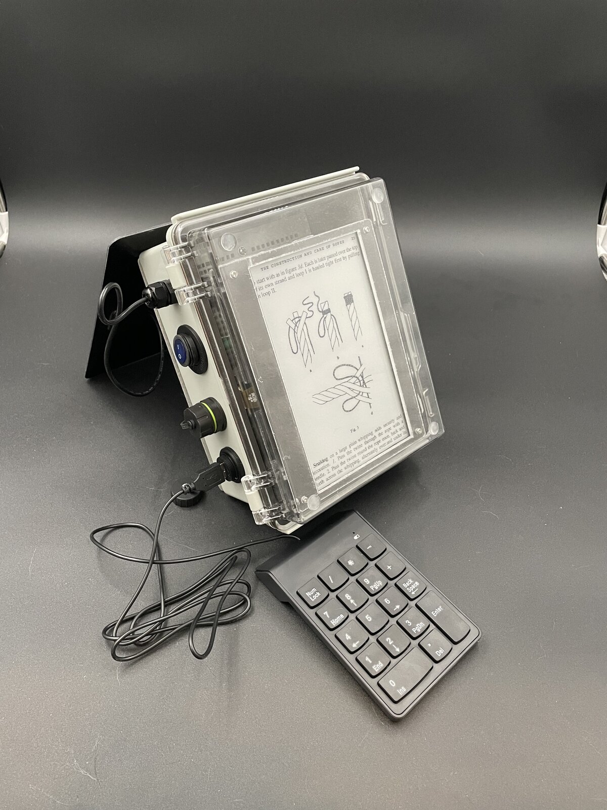 SPRG's e-ink screen showing an illustrated reference page on knot tying, viewed through the clear waterproof lid, with the detachable keypad beside it